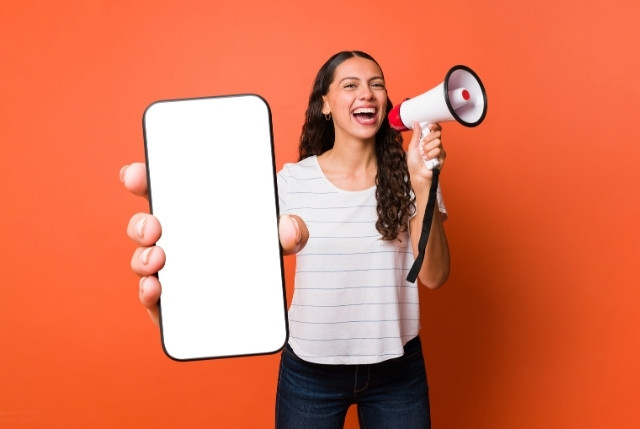 オレンジ色の背景でメガホンを持ちながらスマートフォン画面を見せる女性。日本語学習アプリや外国人社員向けおすすめアプリの紹介をイメージ