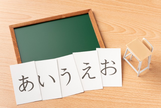 机の上に小さな黒板と椅子の模型が置かれ、その前に『あ・い・う・え・お』と書かれたカードが並ぶ日本語学習のイメージ