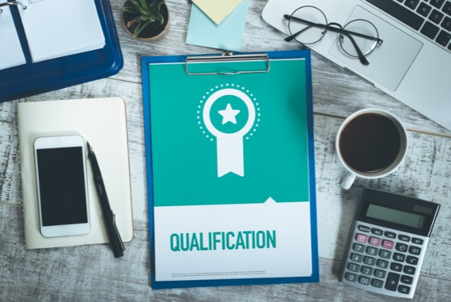 木目調のデスクに資格を示す『QUALIFICATION』と書かれた書類が置かれ、周囲にはノートパソコン、電卓、スマートフォン、コーヒーなどが並ぶ受験資格のイメージ