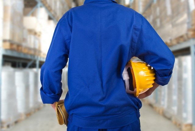 倉庫の棚の前で青い作業服を着た人が、手に黄色いヘルメットを持って立っている後ろ姿