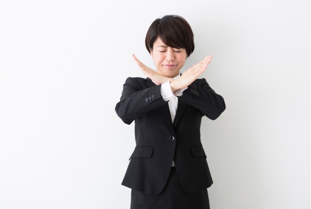 スーツ姿の女性社員が腕でバツ印を作って注意を示す。日本語教育やNG行動の例を伝える場面。
