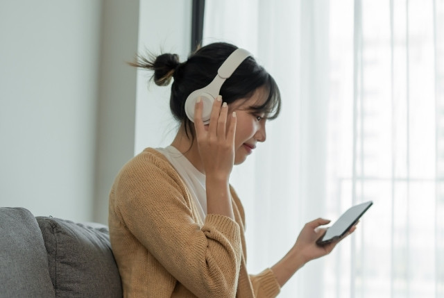 白いヘッドホンで音声を聴きながらスマートフォンを操作する女性。ポッドキャストを楽しむリラックスした室内シーン。