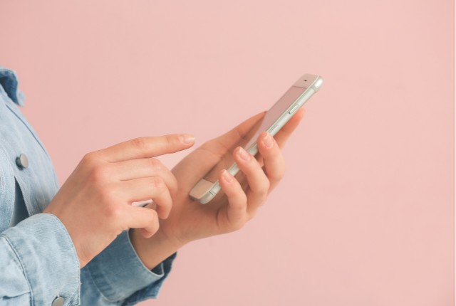 スマートフォンを操作する人の手元の写真。ピンクの背景が柔らかい印象で、eラーニングや語学学習アプリの利用を連想させる。