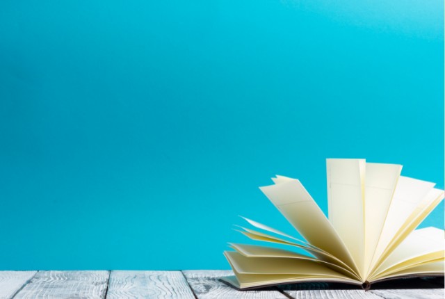 木のテーブルに開いた本が置かれ、明るい青の背景が広がる構図。語彙学習や読書、学びのイメージに最適な写真。