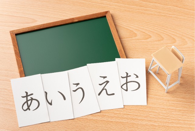 木製の机の上に黒板とミニチュアの机、そして「あいうえお」と書かれたカードが並ぶ日本語学習のイメージ写真