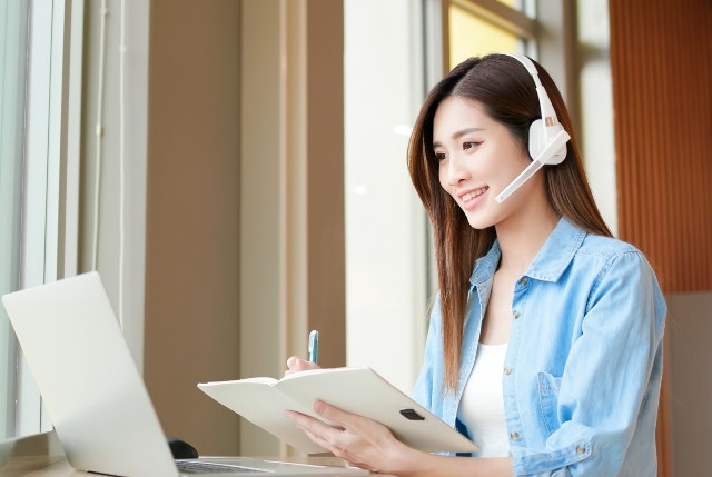 ヘッドセットを着けた女性がノートパソコンを使い、ノートを取りながらオンライン学習を行っている様子。