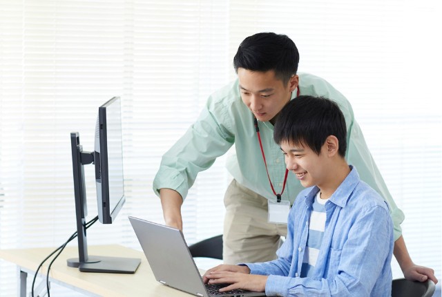 男性社員が若手にパソコン操作を教える様子。職場で日本語指導やコミュニケーション支援を行う場面。