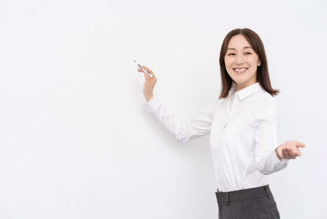 ホワイトボードの前で笑顔で説明する女性講師。日本語教育や教師の指導場面をイメージした写真。