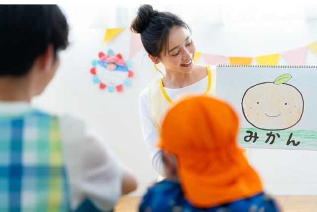 日本語教師が子供に「みかん」の絵を見せて授業を行う様子。ひらがな学習や語彙指導のイメージ。