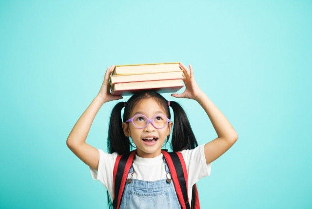 本を頭に乗せて笑う女の子。楽しく学ぶ子供の姿を通して日本語学習への興味や意欲を表すイメージ。