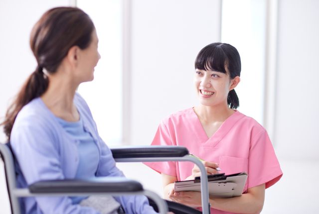 車いす利用者に看護助手が笑顔で対応し、記録を取りながら会話する様子。介護現場のサポート業務のイメージ。
