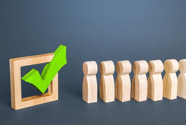 木製の人型フィギュアが一列に並び、左側に緑のチェックマークが入った枠が置かれている様子。外国人介護士の受け入れ体制のイメージ。