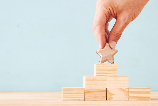 積み重ねた木製ブロックの頂点に星型ブロックを置く手のイメージ。努力や成長、目標達成を象徴する場面。