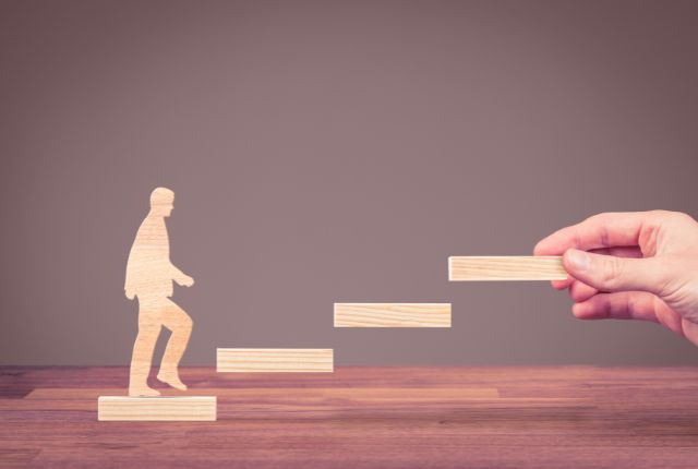 木製の人型が階段を一歩ずつ上る様子を描いた画像。企業の人材育成やキャリアステップ支援の成長を象徴。