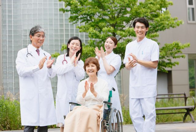 医師と看護スタッフが車いすの高齢者を囲んで笑顔で拍手する様子。介護・医療現場の支援イメージ。