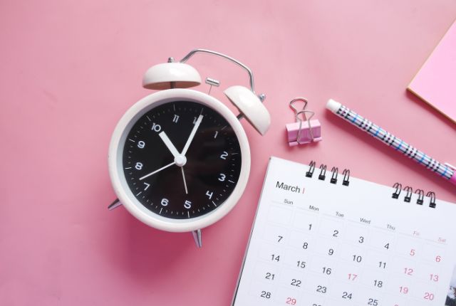 淡いピンクの背景に白い目覚まし時計と3月のカレンダー。日本語能力試験直前の時間管理と準備をイメージ。