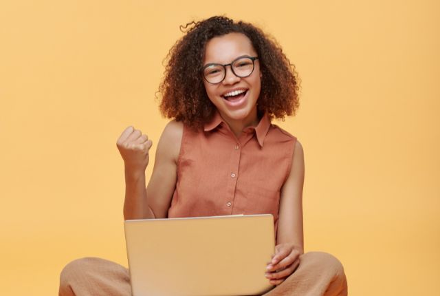 ノートパソコンで学習し笑顔でガッツポーズをする女性。オンラインでスキルアップや学習成果を喜ぶ様子を表現。