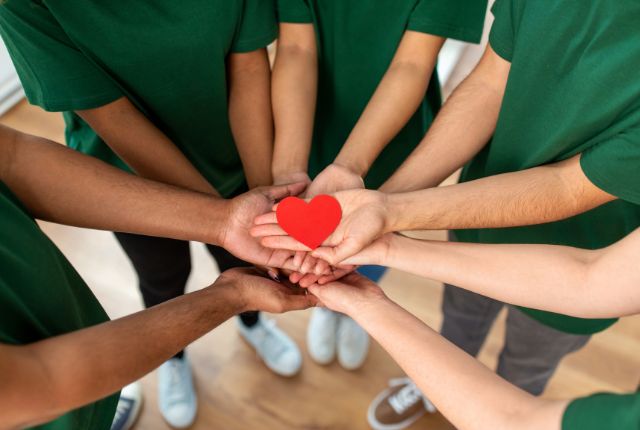 緑のTシャツを着た人々が円になり、手のひらに赤いハートを乗せて支え合う様子。介護や支援のチームワークを表すイメージ。