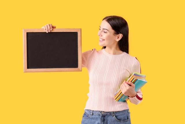 笑顔の外国人女性が黒板を手に持ち、もう片方の手に色とりどりのノートを抱えている。日本語教授法の学びを象徴するシーン。