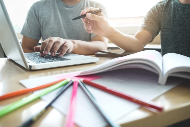 ノートパソコンとノートを使って学習する外国人と教師。ペンを手に協力しながら勉強を進める様子。