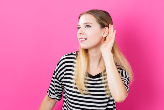 耳に手を当てて聞こうとしている人物。ピンク背景で聴解や注意深く聞く姿勢をイメージさせる写真。