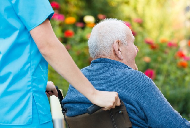 車椅子の高齢者を介護士がサポートしながら屋外散歩する様子。介護や福祉サービスのイメージ。