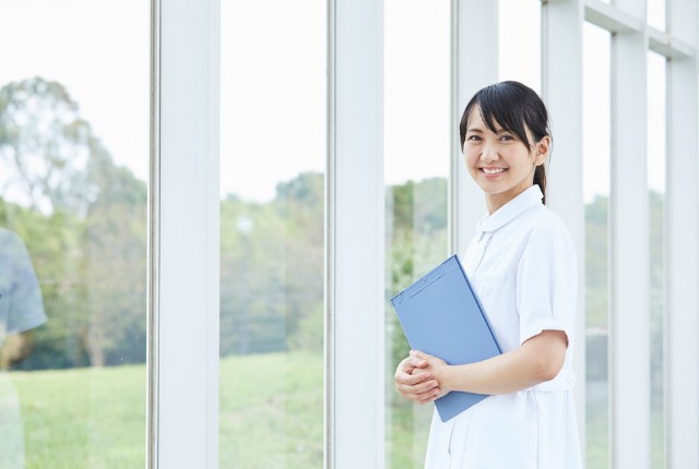 窓辺で書類を持ち笑顔を見せる介護士。医療や介護の現場で働くスタッフのイメージ。