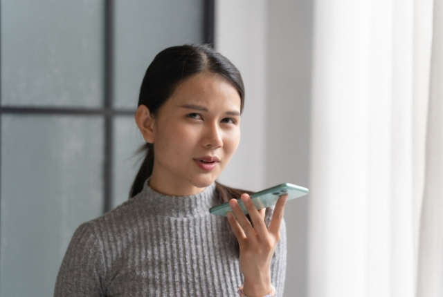 室内でスマートフォンに向かって話しかける若い女性のイメージ写真