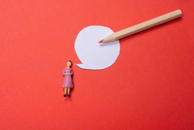 赤い背景に白い吹き出しと鉛筆、小さな女性人形が置かれたイメージ