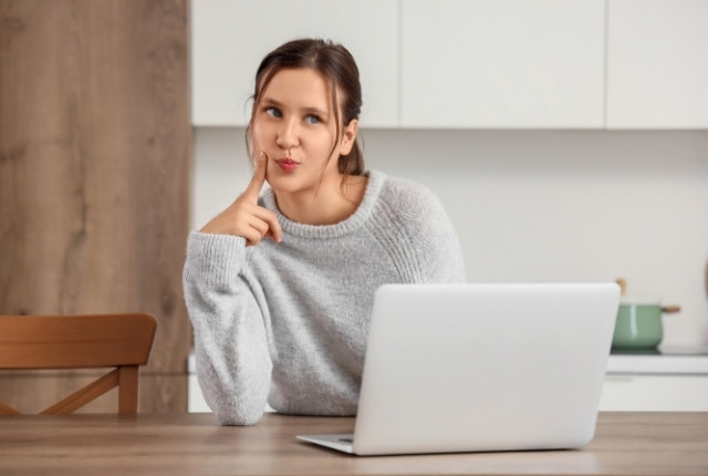 キッチンのテーブルでノートPCを前に、指を頬に当てて考え込む女性