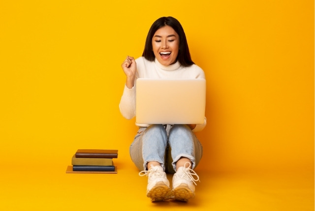 床に座りノートパソコンを操作しながら喜ぶ表情を見せる人物と本の束